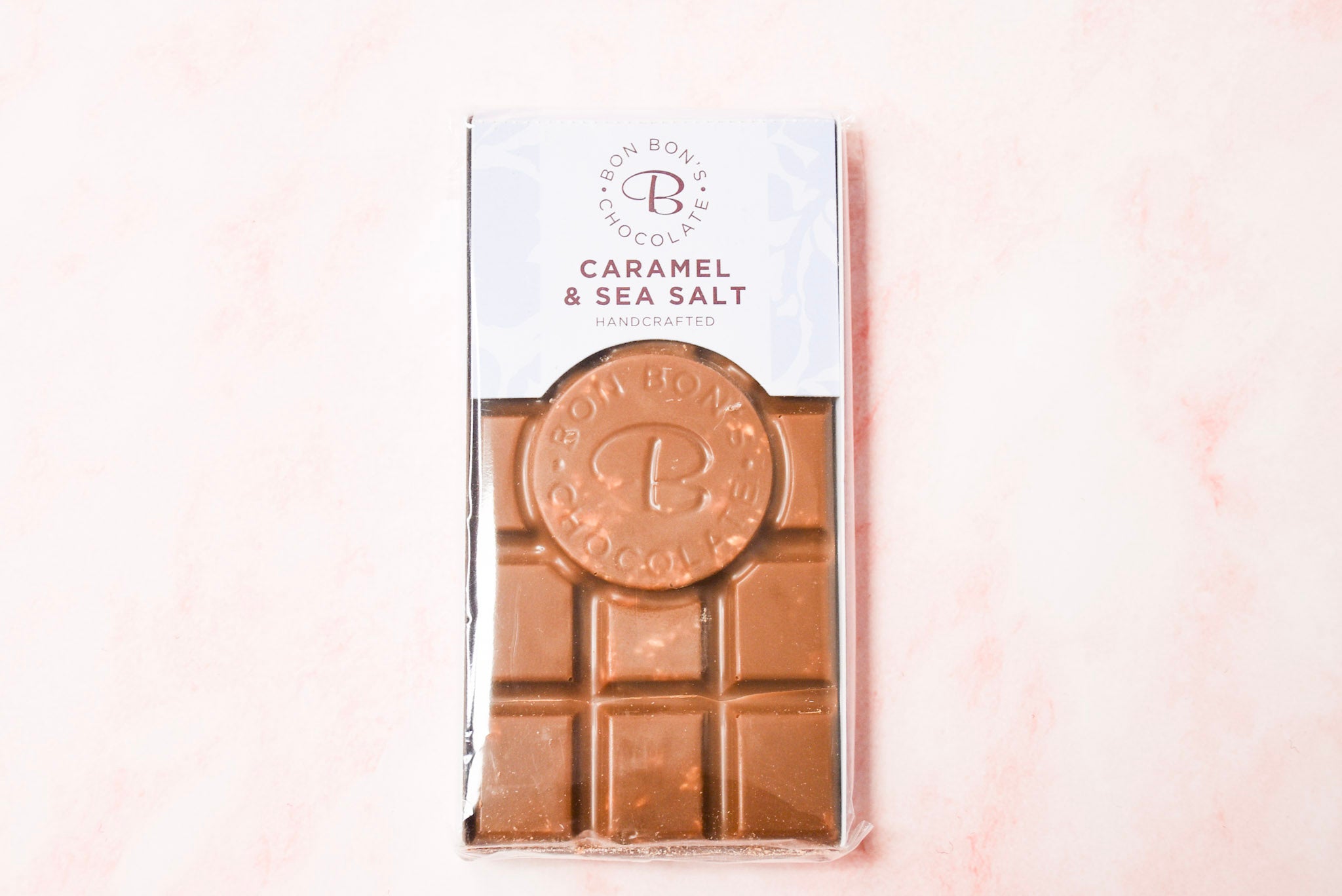 This image features a luxurious Bon Bon's Handcrafted Caramel & Sea Salt Chocolate Bar, presented in an elegant packaging with floral patterns. The chocolate bar itself is visible through a clear window, displaying its rich, layered texture, the bar promises a caramel & sea salt chocolate experience.