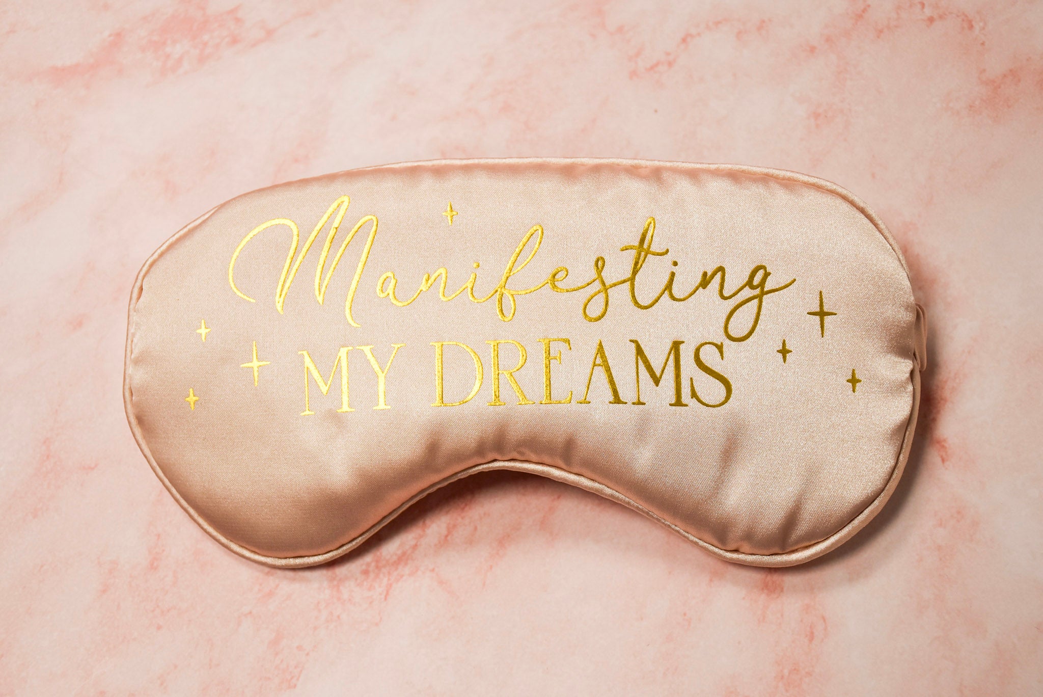 A pink silk eye mask with the text 'Manifesting My Dreams' written in a golden script.