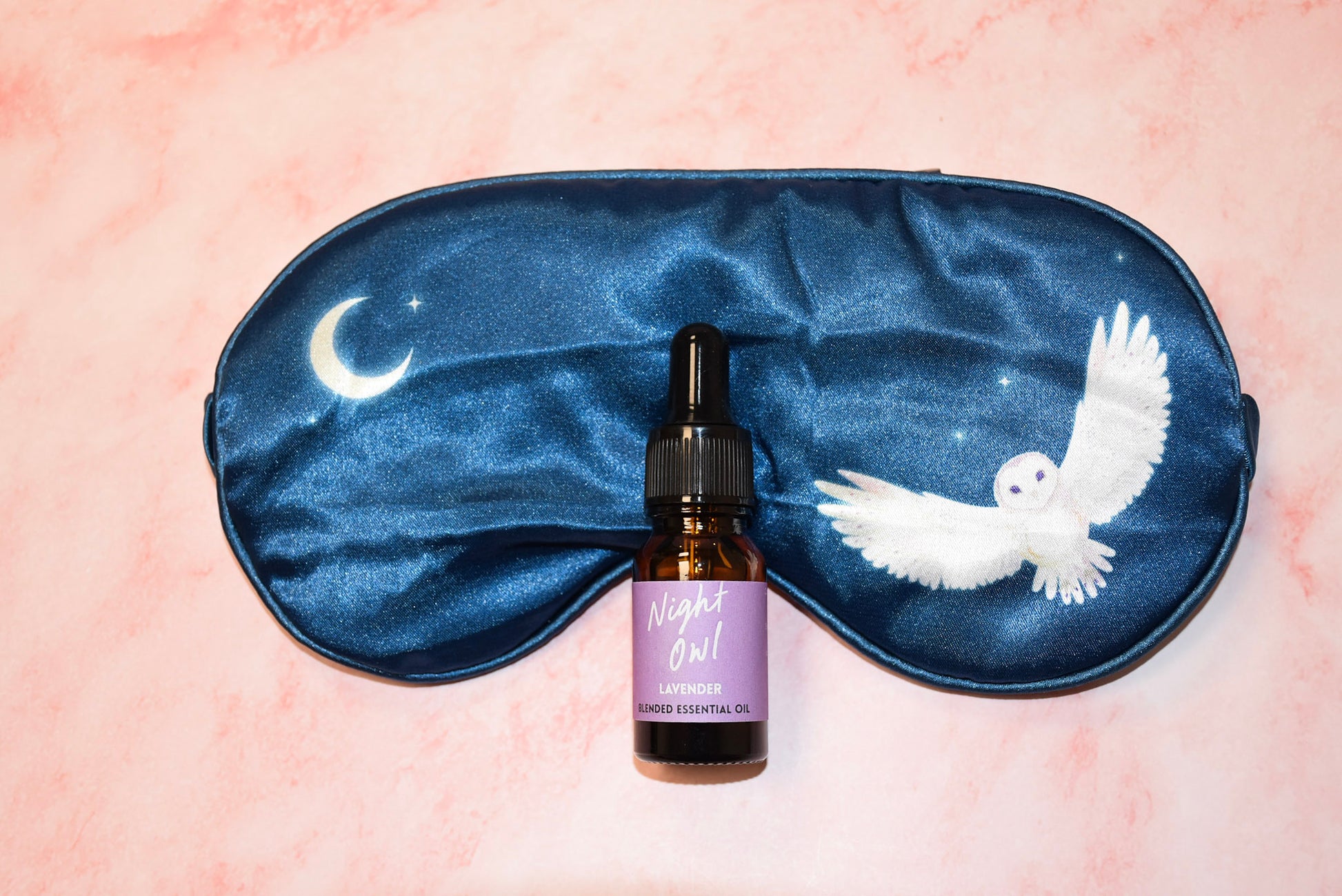 A blue silk eye mask featuring an owl design, accompanied by a small bottle of lavender oil.